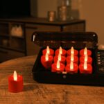 Tolv röda uppladdningsbara LED-värmeljus laddas i en svart laddbox på ett träbord, ett rött ljus ligger isär. Ett stämningsfullt, svagt upplyst vardagsrum syns i bakgrunden.