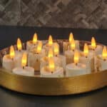 Ett runt guldfärgat serveringsfat med flera LED-värmeljus med 3D-flamma, uppladdningsbara, med timerfunktion och fjärrkontroll, arrangerade på en mörk bänkskiva med en kaklad vägg med struktur i bakgrunden.