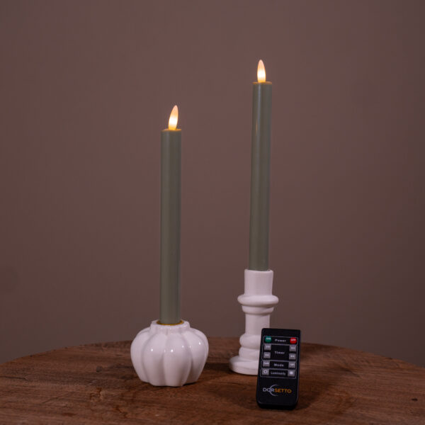 Två tända LED-middagsljus 400 timmars brinntid, uppladdningsbara, olivgröna (set om 2) står i vita keramikhållare på en träyta, med en liten svart Doggetto-fjärrkontroll framför. Bakgrunden är enfärgat brun.