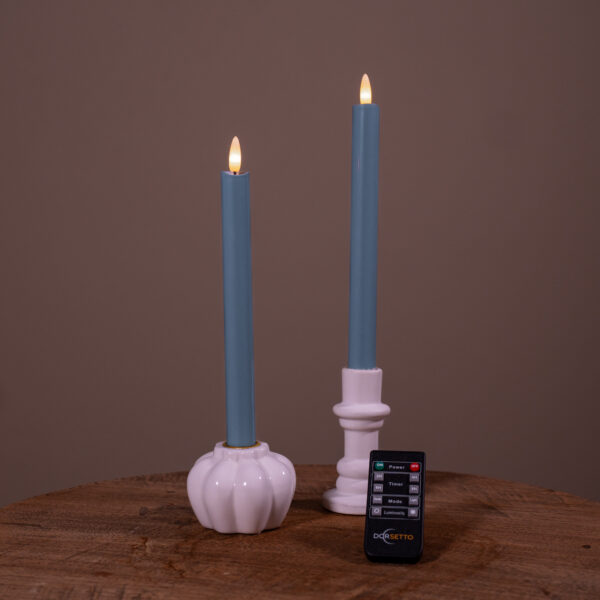 Två stenblå LED-dinerljus (set om 2, upp till 400 timmars brinntid) i vita hållare på en träyta, med svart fjärrkontroll framför och enfärgad brun bakgrund.