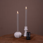 Två mörkgrå LED-dinerljus (set om 2, uppladdningsbara, upp till 400 timmars brinntid) i vita hållare på en träyta med brun bakgrund och en liten Dorsetto-fjärrkontroll framför.