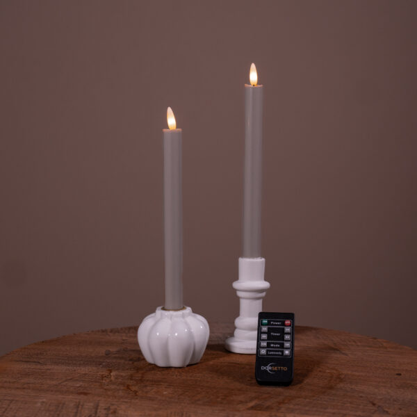 Två mörkgrå LED-dinerljus (set om 2, uppladdningsbara, upp till 400 timmars brinntid) i vita hållare på en träyta med brun bakgrund och en liten Dorsetto-fjärrkontroll framför.