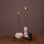 Två beige LED-dinerljus (set om 2, 400 timmars uppladdningsbar brinntid) står i vita hållare på en träyta, med en liten svart fjärrkontroll framför. Den enfärgade bruna bakgrunden kompletterar scenen.