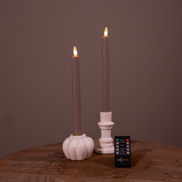 Två beige LED-dinerljus (set om 2, 400 timmars uppladdningsbar brinntid) står i vita hållare på en träyta, med en liten svart fjärrkontroll framför. Den enfärgade bruna bakgrunden kompletterar scenen.