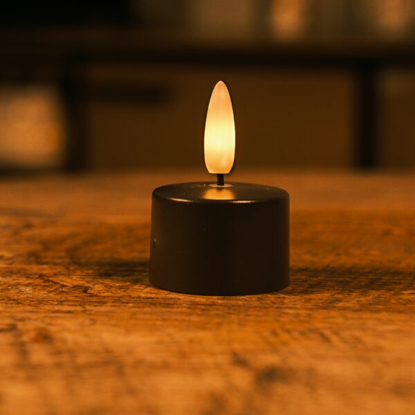 Ett litet batteridrivet LED-ljus med varmt, fladdrande sken placerat på en träyta och skapar en mysig atmosfär, med mjukt suddig bakgrund.