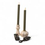 Två gröna LED-ljus med cremefärgade hållare, en svart fjärrkontroll och en svart nätadapter står tillsammans mot en vit bakgrund. Ljusen har flammformade toppar.