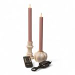 Två mjukt rosa LED-dinerljus i vita hållare tillsammans med fjärrkontroll och svart nätadapter mot vit bakgrund. Ljusen har realistiska flamformade toppar.